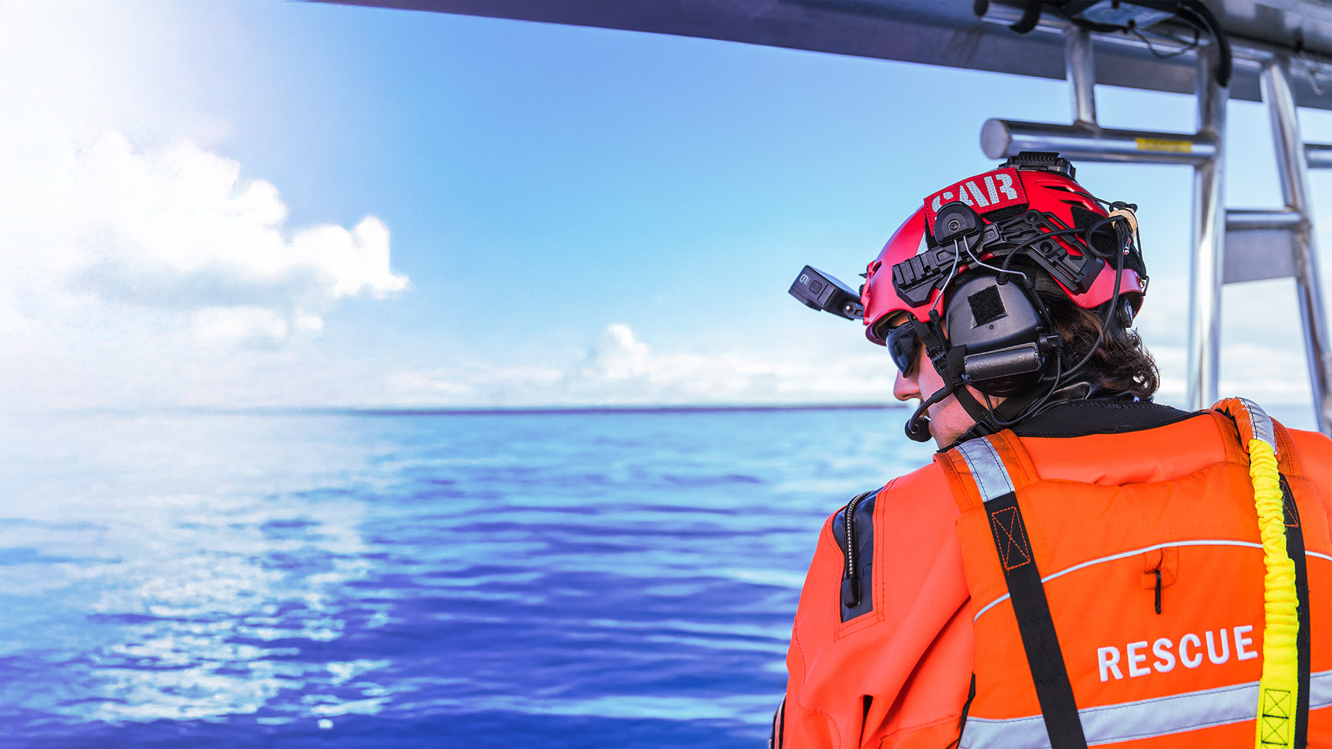 Image of man wearing an orange jacket and red Team Wendy helmet with a SAR patch, comms unit and headlamp on the water