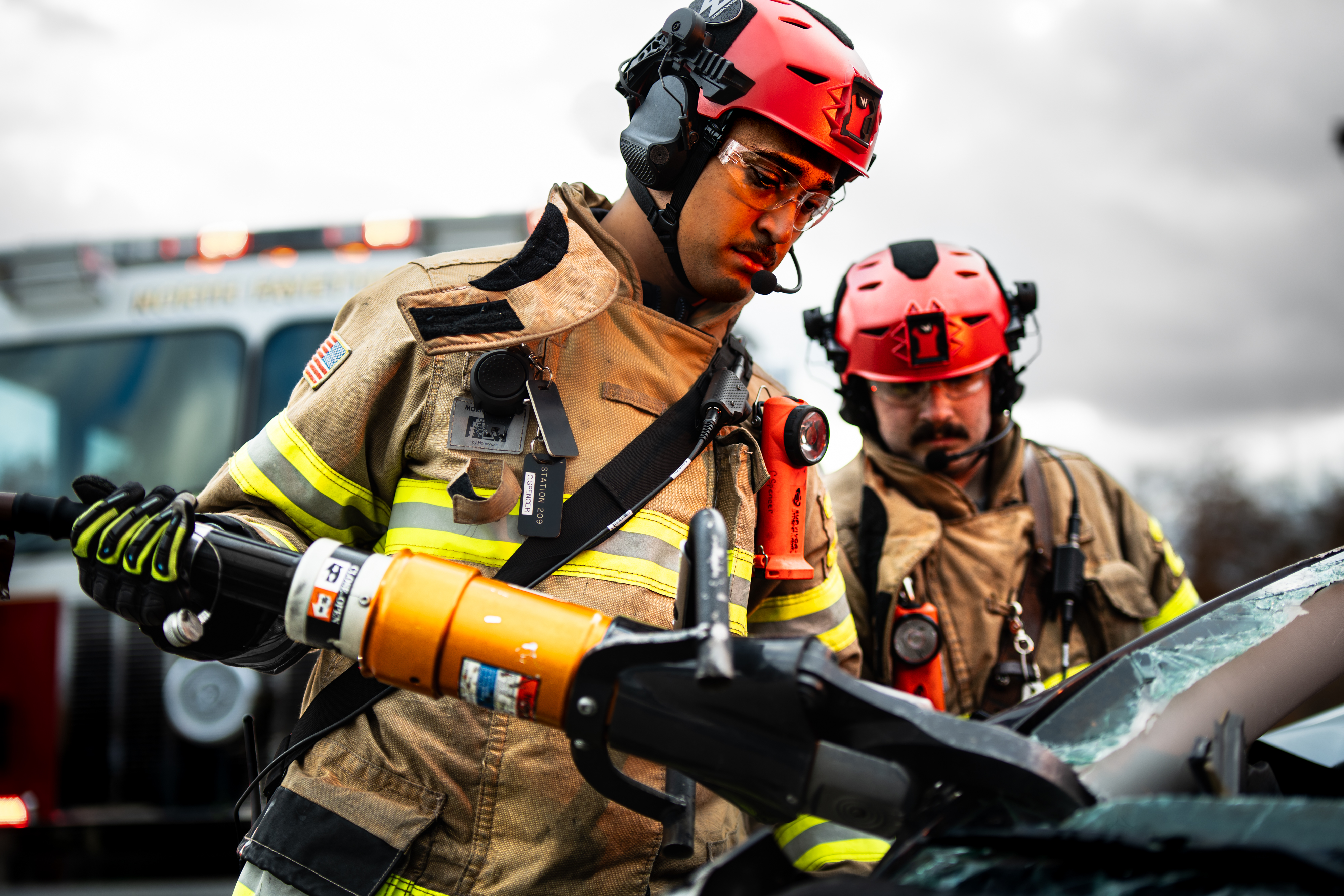 Two firefighters wearing tan turnout gear and red Team Wendy EXFIL Rescuer helmets are performing a vehicle extrication. One firefighter is operating a hydraulic rescue tool (commonly known as the Jaws of Life) to cut through the damaged vehicle, while the other stands nearby assisting.